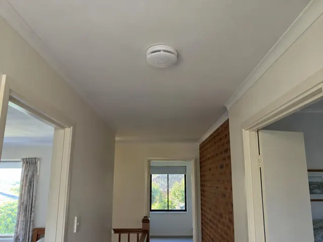 Ceiling-mounted smoke alarm in a hallway with doorways and a brick feature wall in Wadalba